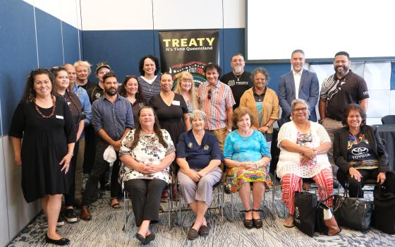 A group of ITTB Board members and Community members gathered together smiling with a Truth and Treaty banner in the background